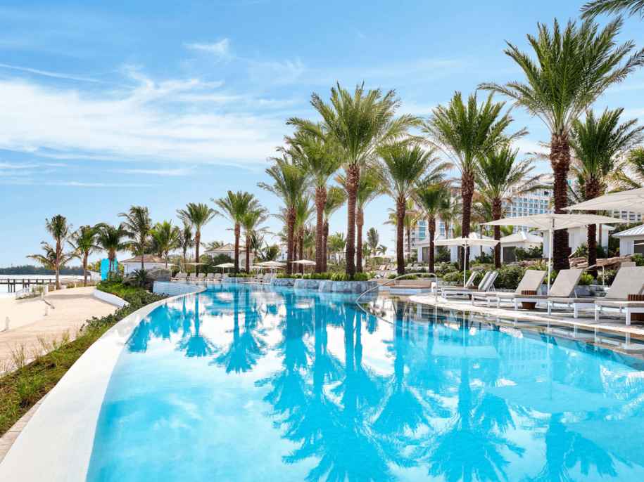 Imagen de una gran piscina con palmeras y tumbonas con vistas a la playa. 