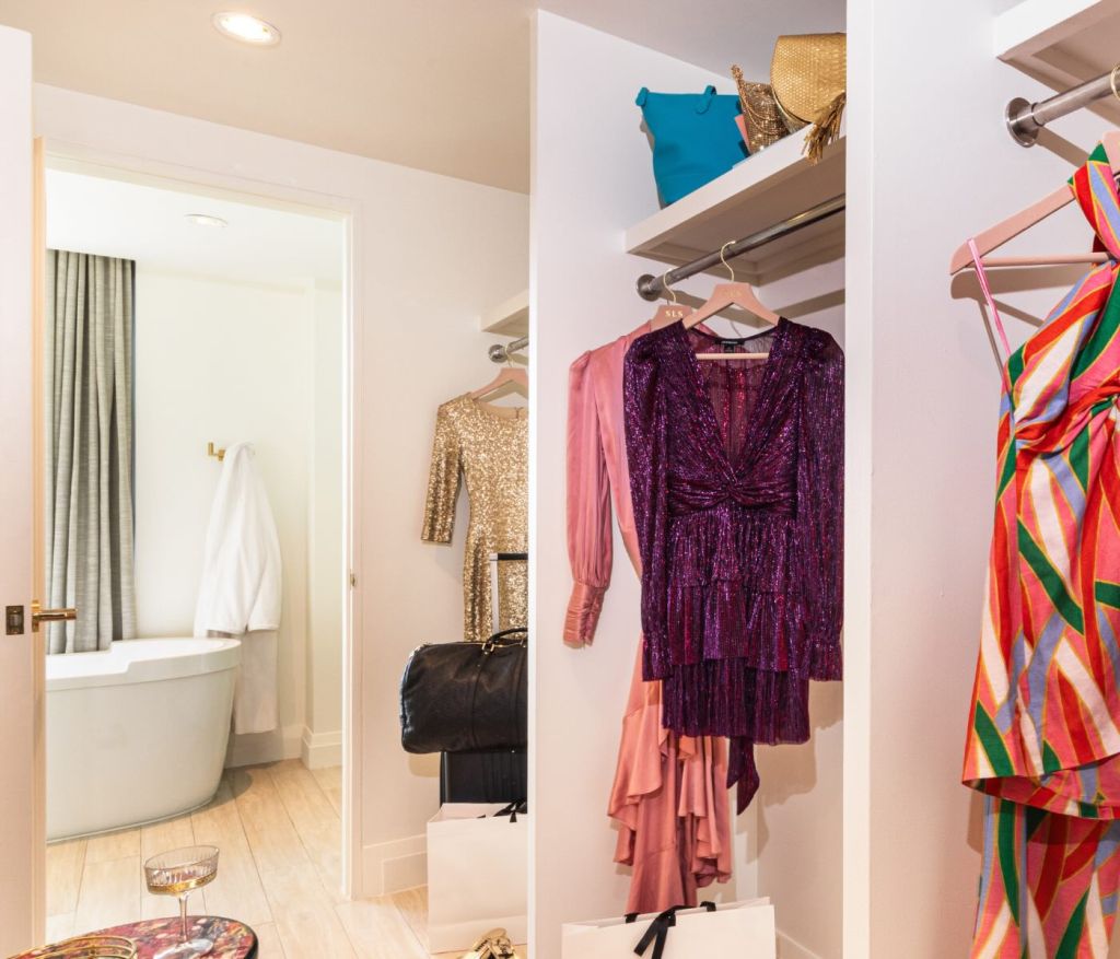 a closet full of elegant clothes with a door through to the bathroom with a large tub