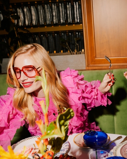 a woman in a feather cap tasting food next to a woman in a bright pink top both sitting in a velvet green booth