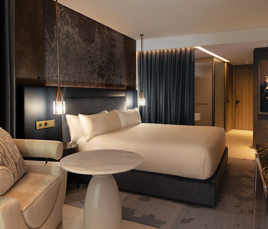 A hotel room with an accent table and arm chair in the foreground, and a king size bed, with decorative headboard, flanked by hanging bedside lights in the background