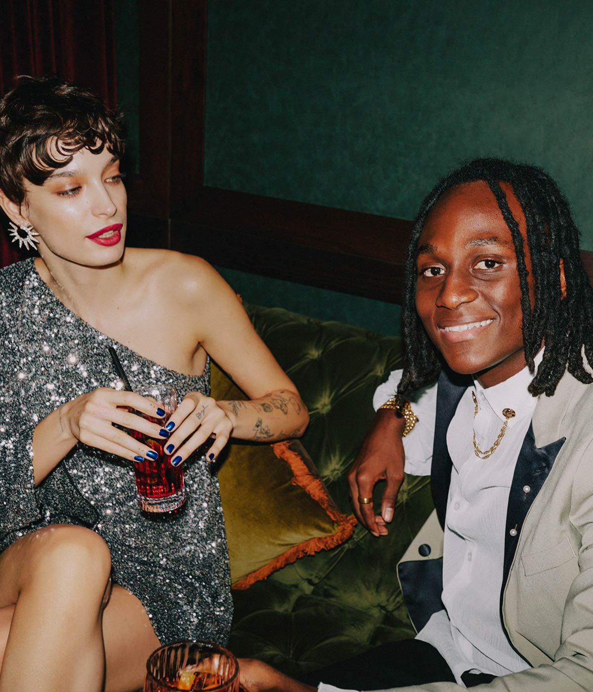 A woman in a sparkly cocktail dress, holding a cocktail and looking at a man who is looking at the camera smiling