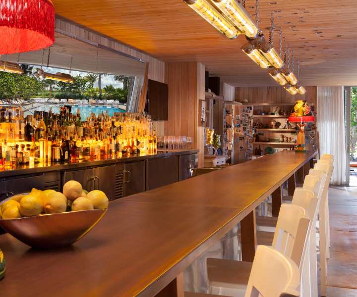 Bar de madeira comprido, totalmente abastecido, com uma taça de limões e cadeiras altas brancas. 