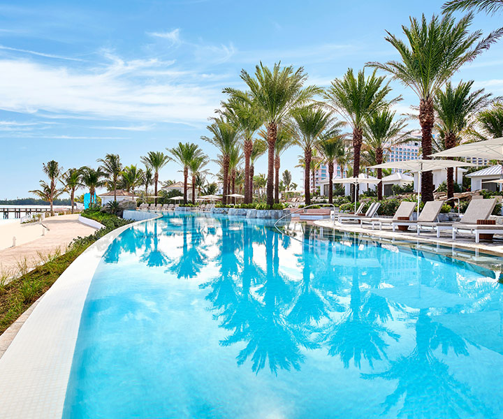 Imagen de una gran piscina con palmeras y tumbonas con vistas a la playa.