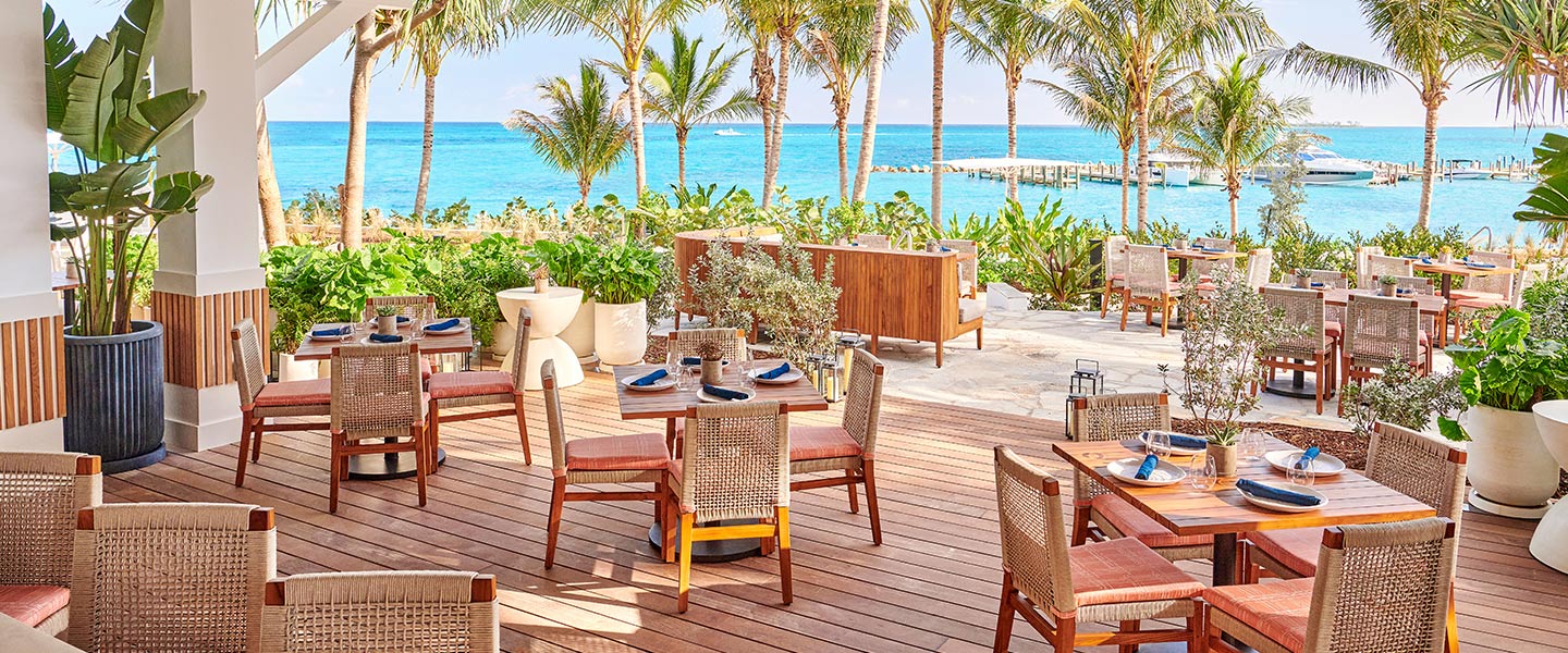Mesas e cadeiras de madeira no terraço de um bar à beira-mar com vista para o oceano.