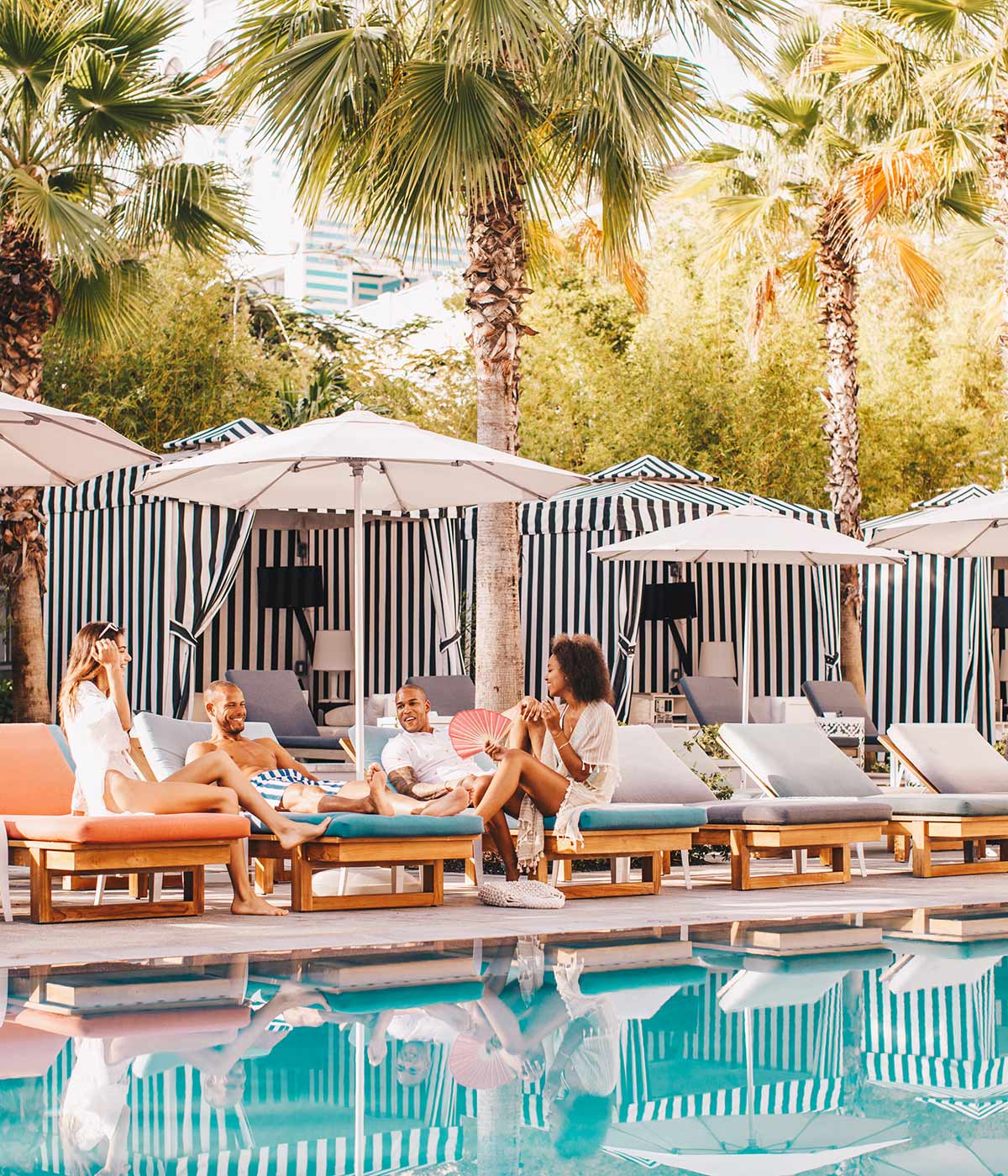 group of friends sitting and talking on loungers by the pool, in front of a row of striped cabanas