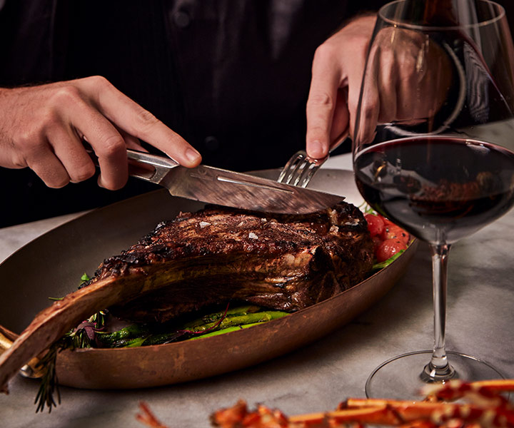 Uma pessoa a cortar um bife tomahawk sobre espargos, com um copo de vinho tinto.