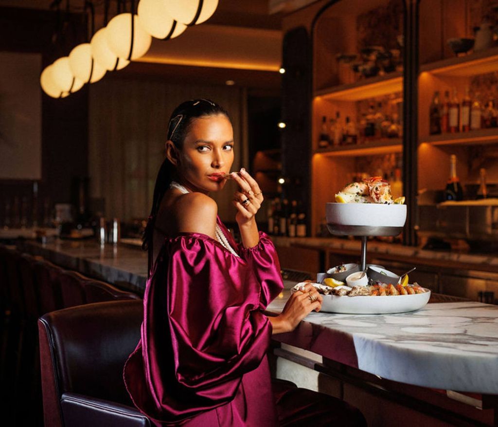 Mulher com um vestido drapeado de seda magenta a comer um prato de marisco no bar e a olhar por cima do ombro