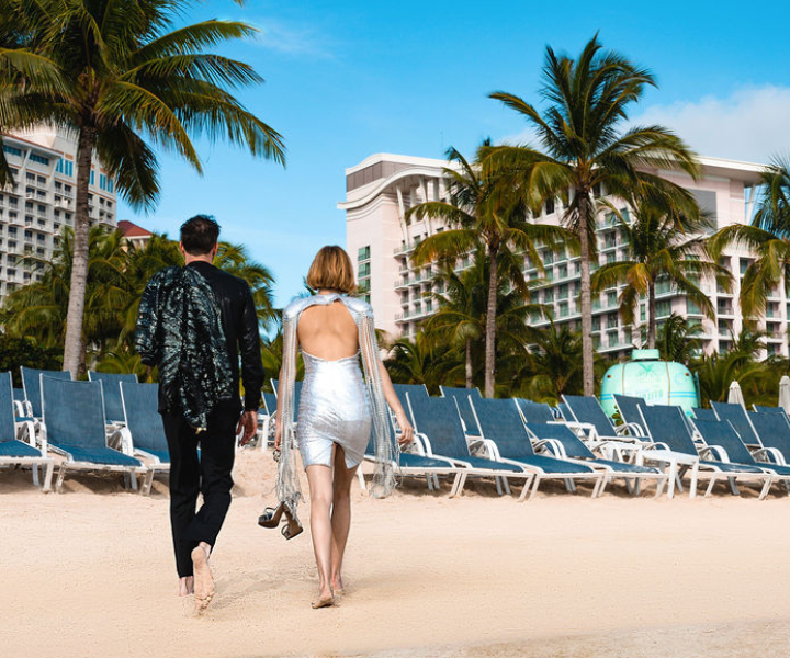 a woman in a silver dress and a man in a black suit walking on the beach lined with lounge chairs