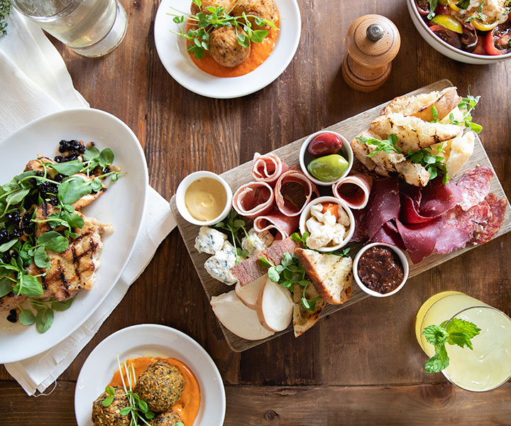 A rustic table laid with various dishes including charcuterie, sandwiches, salads, and drinks.