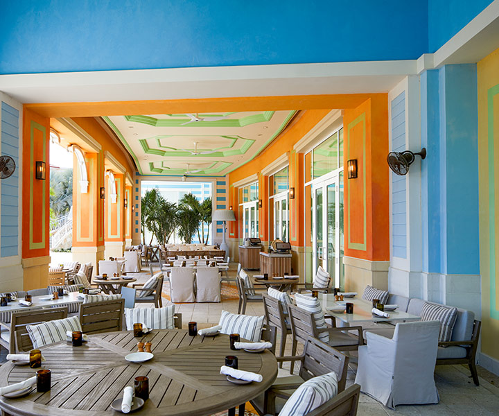 Área de refeições ao ar livre no restaurante Fi'lia com paredes coloridas, mesas e cadeiras de madeira, com vista para a vegetação tropical.