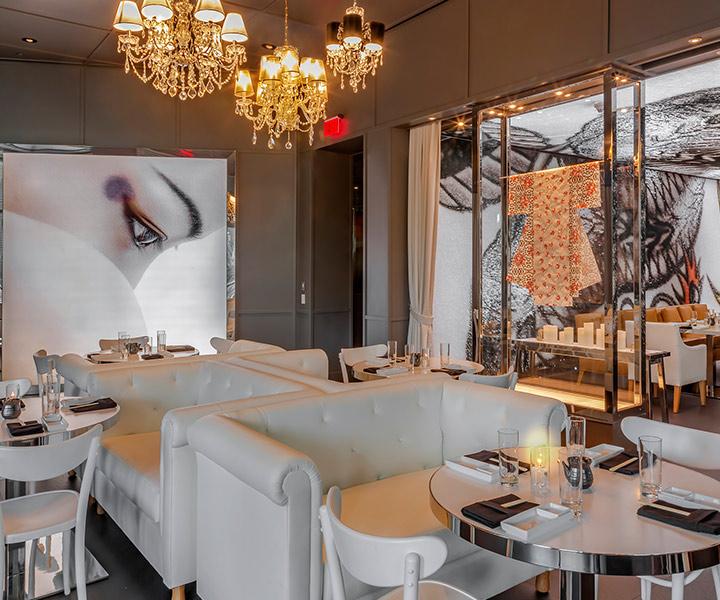 Intérieur du restaurant Katsuya Baha Mar, avec des tables et des chaises blanches, un lustre en cristal et une peinture murale du visage d’une femme sur le mur.