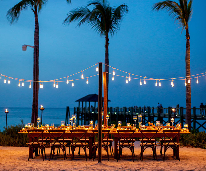 Mesa larga en la playa con velas y guirnaldas de luces creando un ambiente acogedor.