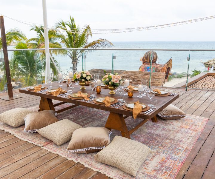 Mesa baja preparada para un evento con cojines como asientos, en una terraza con vistas al mar. 