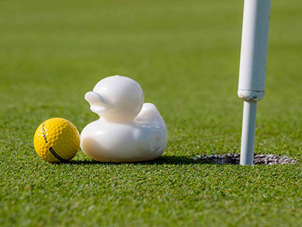 Close-up of a putting green with a white rubber duck and golf ball next to the golf hole with a pin.