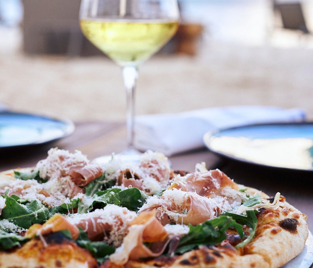 Pizza topped with prosciutto with a crisp glass of white wine in the background