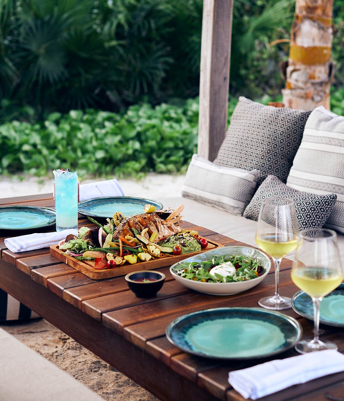 Table in the sand, surrounded by seating, full of fresh food, salad and white wine