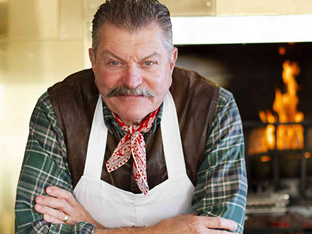 Portrait of Celebrity Butcher Dario Cecchini, a man with a mustache wearing a chef's apron, folding his hands in front of a fireplace.