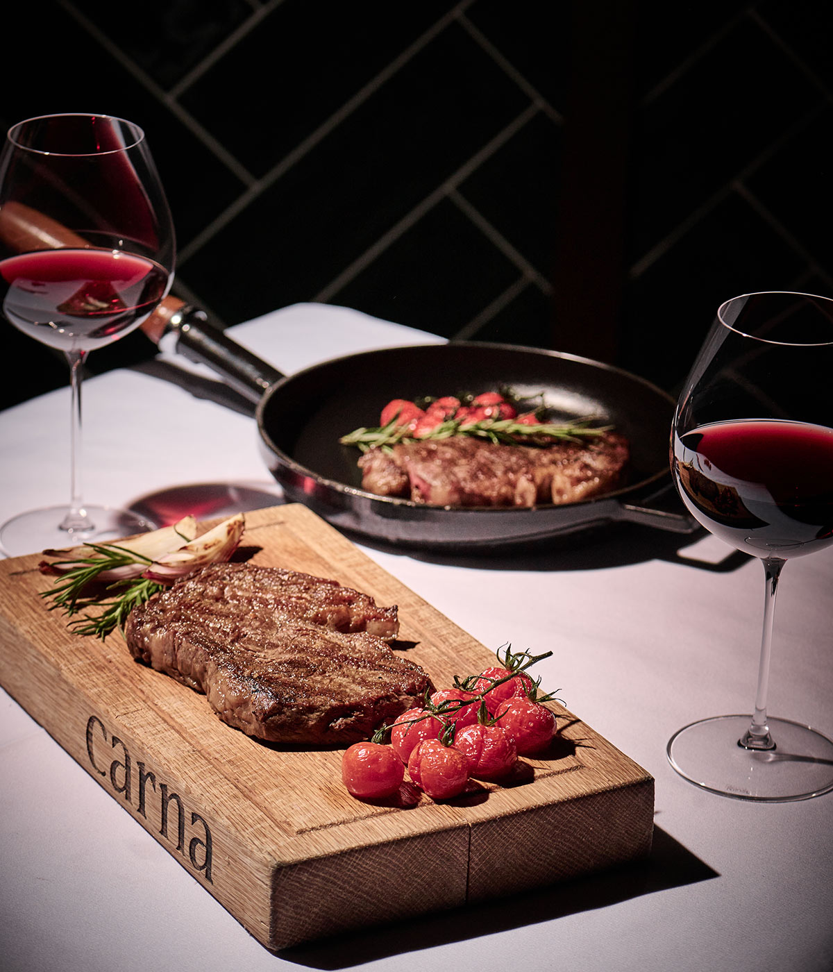 Bife cozinhado numa tábua de cortar Carna, ladeado por dois copos de vinho tinto. 