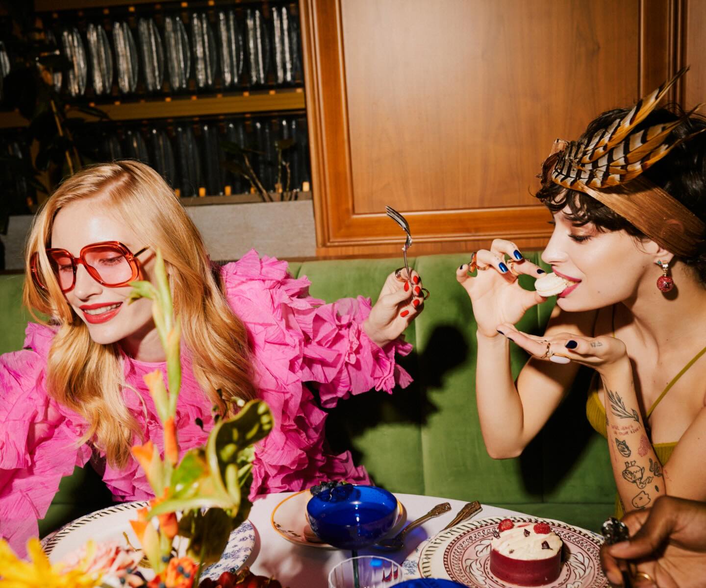 a woman in a feather cap tasting food next to a woman in a bright pink top both sitting in a velvet green booth