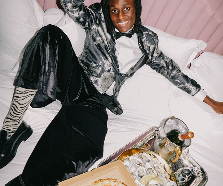 A sophisticated man in a tailored suit and tie, elegantly seated on a luxurious bed with a tray of gourmet food.