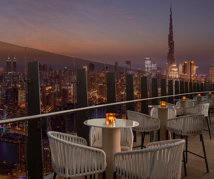 Balcony with tables, overlooking Dubai skyline illuminated at night with twinkling lights and tall buildings including the Burj Khalifa. 