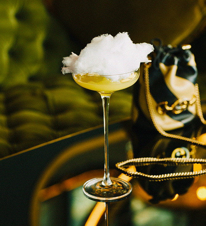 Impressionante cocktail amarelo com um topo de algodão doce, assente numa mesa espelhada com uma bolsa de designer em segundo plano.