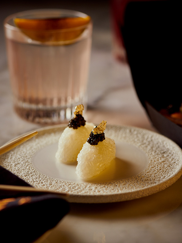 Dos piezas ornamentadas de sushi decoradas con caviar y copos de oro en un plato redondo y blanco y un cóctel.