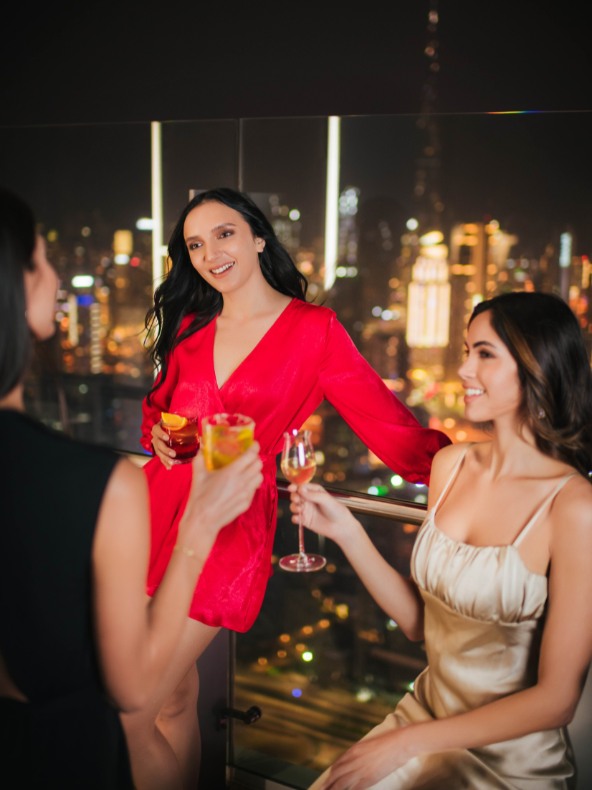 Two women holding cocktails and smiling with the Dubai skyline behind them at night