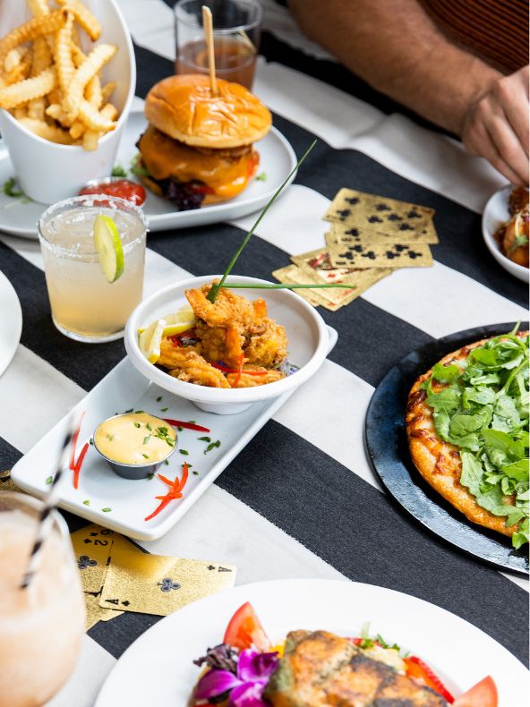 una mesa repleta de comida, con hamburguesas, cócteles, patatas fritas y pizza