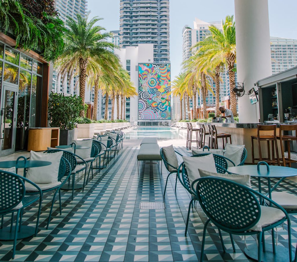 Pátio no rooftop com cadeiras e mesas, orlado de palmeiras luxuriantes e com um mural colorido junto à piscina.