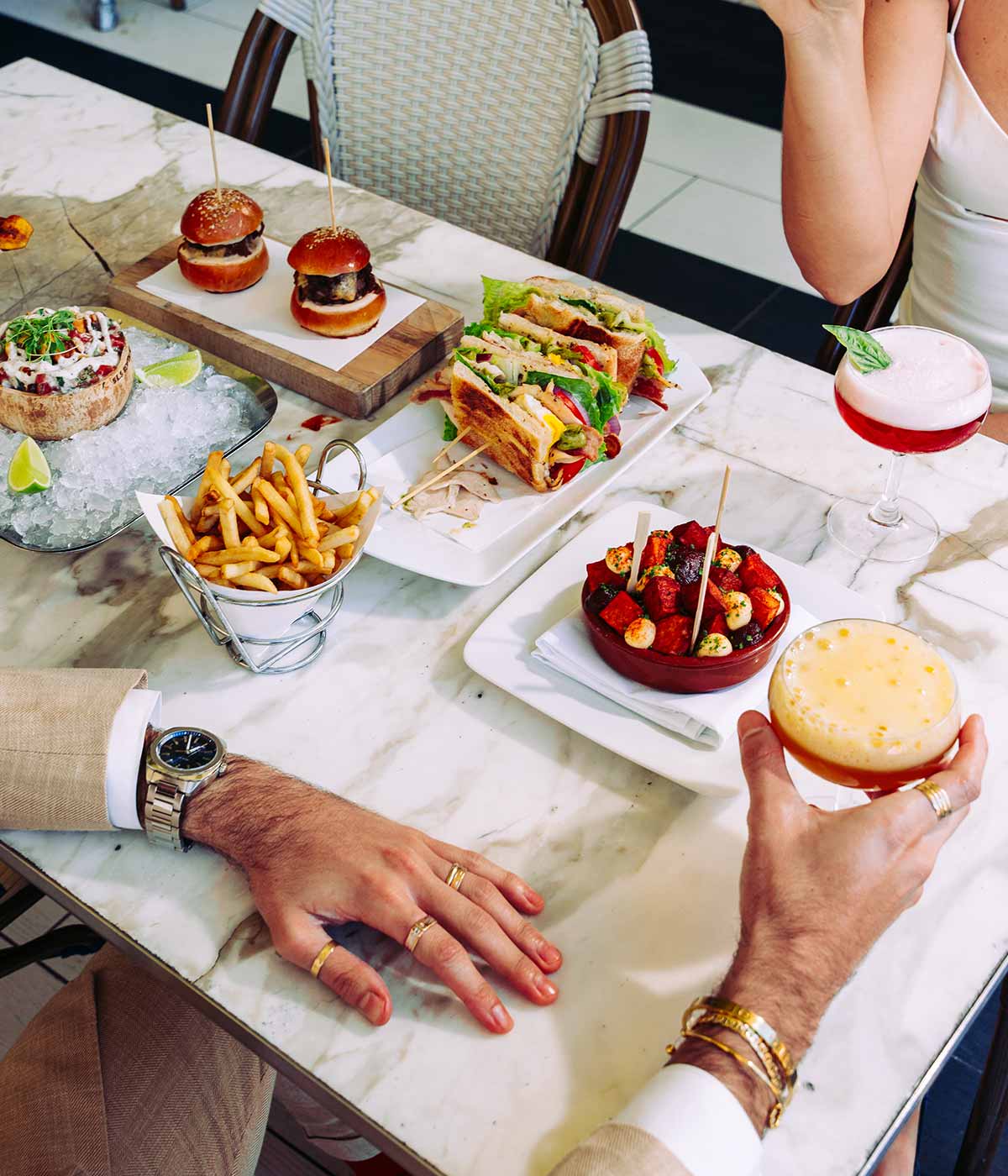A spread of food including french fries, sandwiches and cocktails, being eaten by two people