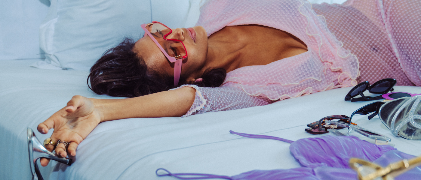 a woman in rose colored glasses and a pink dress lounging on a king bed holding onto silver heels
