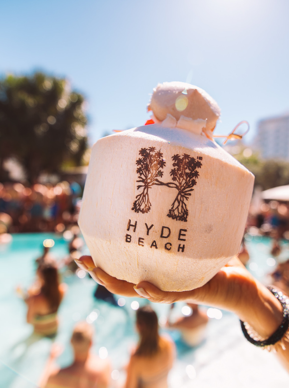 a beverage within a coconut enscribed with Hyde Beach being held up in front of the pool