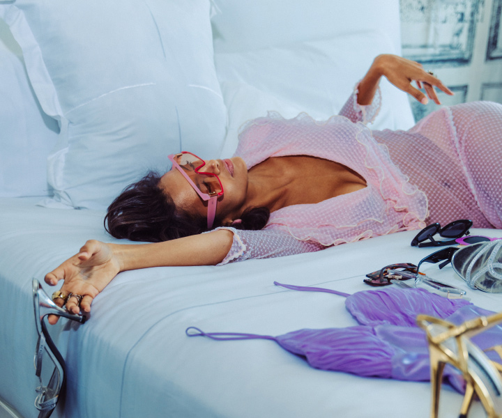 Femme élégante allongée sur un lit luxueux, entourée d'oreillers moelleux, de draps, de vêtements et de chaussures.