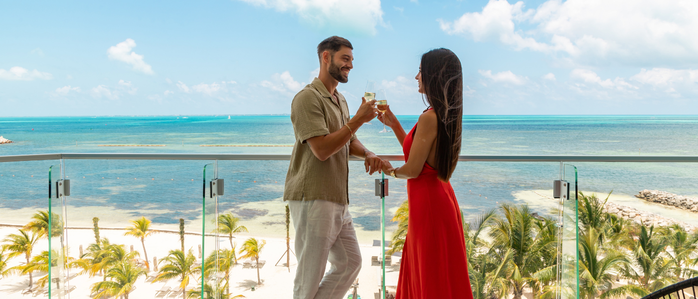 a woman in a long red dress is holding champagne and smiling at her partner who is also holding champage. Both are standing on a large terrace that overlooks the ocean