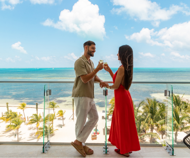 a woman in a long red dress is holding champagne and smiling at her partner who is also holding champage. Both are standing on a large terrace that overlooks the ocean