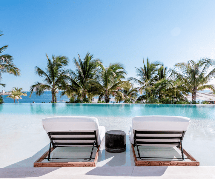 two luxurious lounge chairs sitting next to a beach entrance style infinity pool facing palm trees