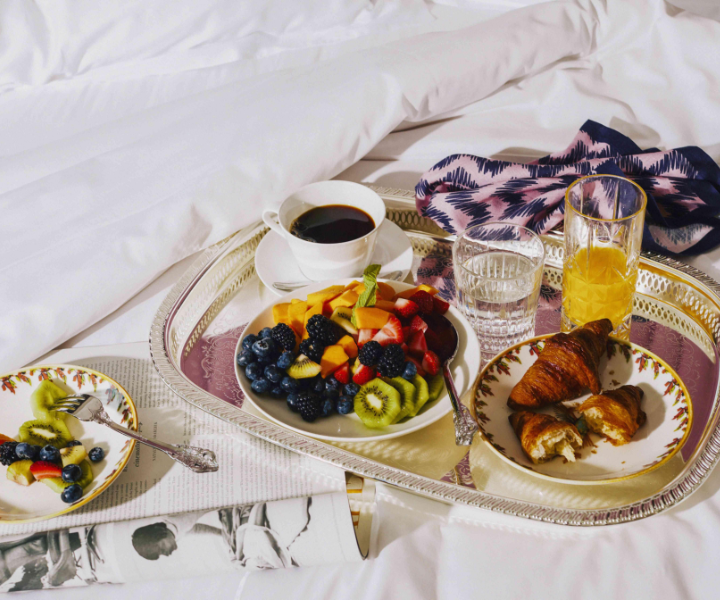 Una escena lujosa con una taza de café, un delicioso desayuno y una revista elegantemente colocada en una lujosa cama, creando un ambiente acogedor y sofisticado.