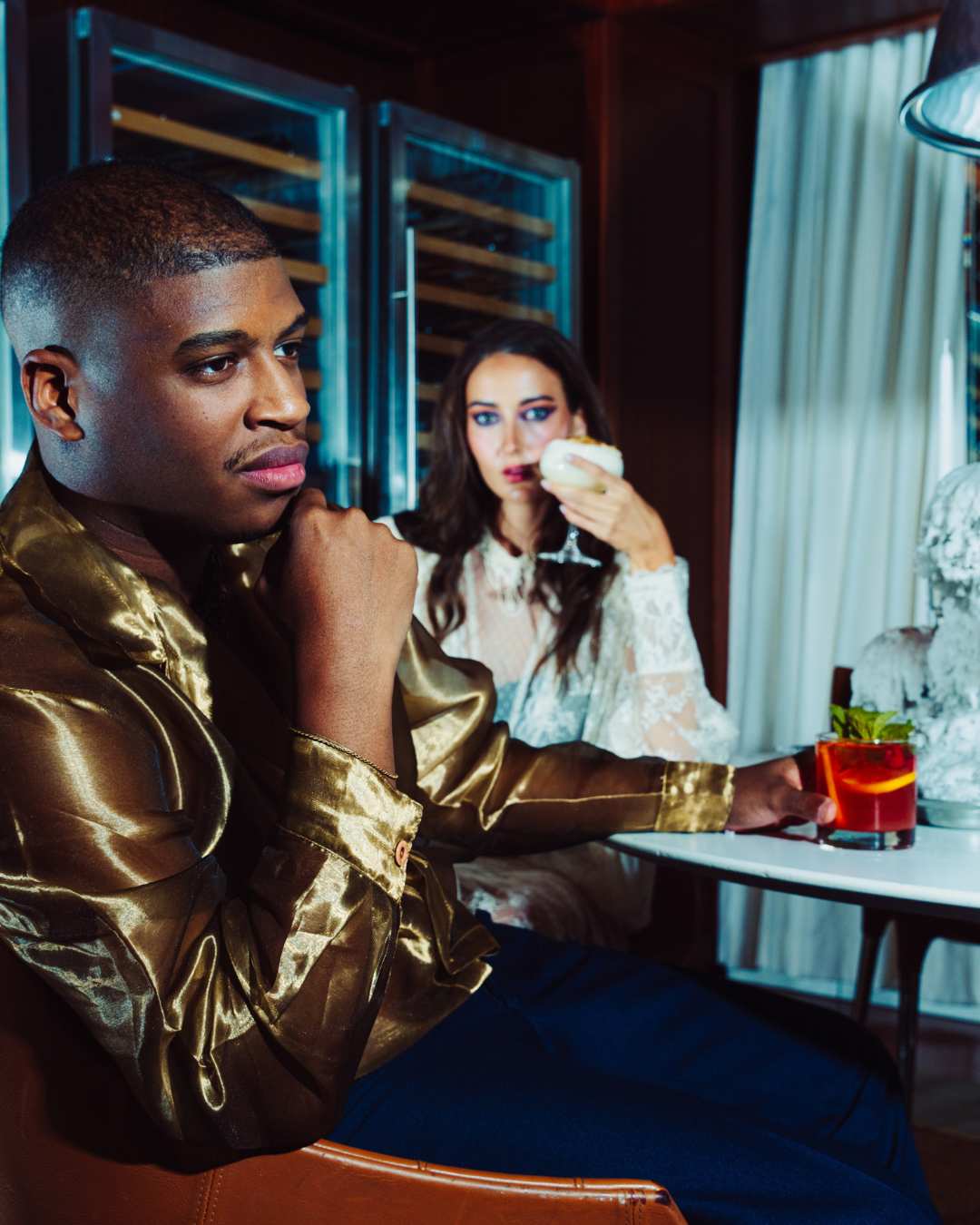 a male and female model sit at a table in high fashion outfits with cocktails in front of them