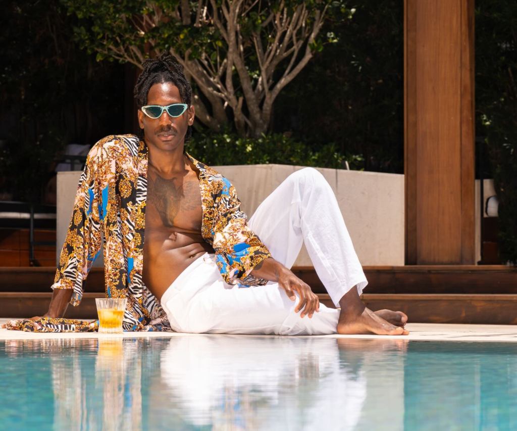 a man in stylish swim cover ups sitting by a pool with a cocktail in hand