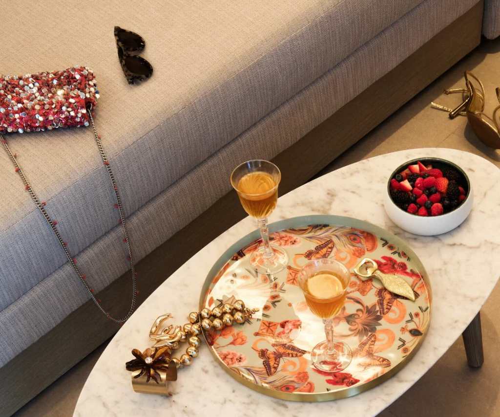 a colorful platter with two glasses of rosé and a bowl of berries next to it on the coffee table as well as large gold jewlry.