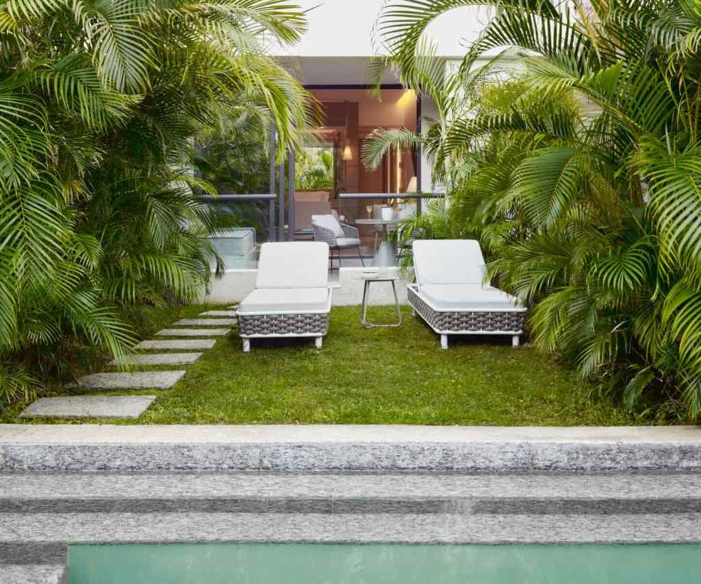 two lounge chairs on grass in front of a room with a deck, and stairs in front of them leading to a pool