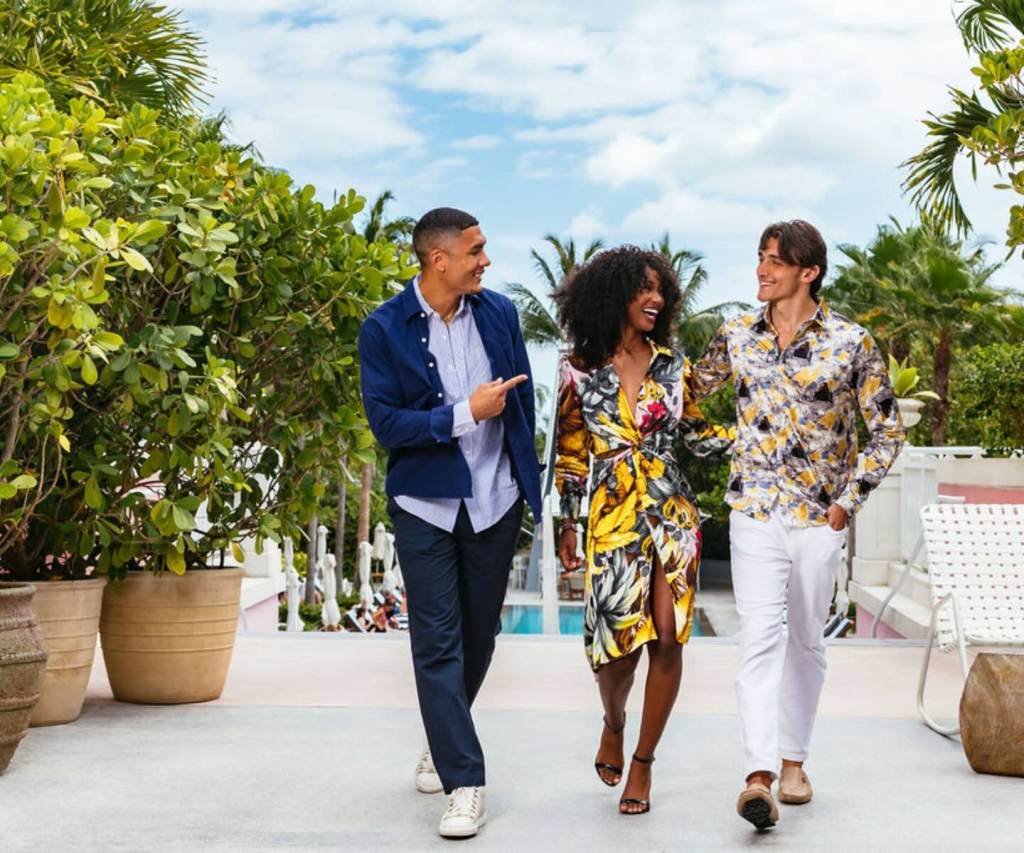 two men and a woman walking outside SLS Baha Mar with a pool in the background, surrounded by beautiful and lush greenery