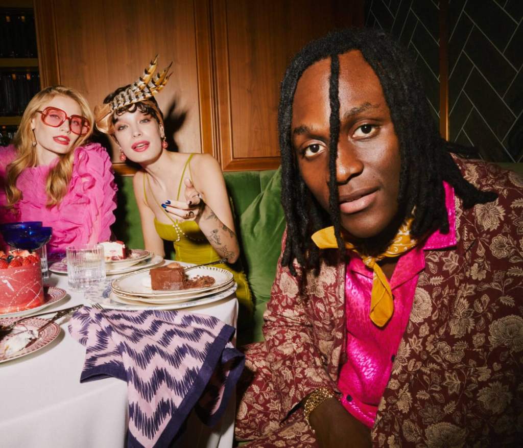 a woman in a feather cap tasting food next to a woman in a bright pink top both sitting in a velvet green booth
