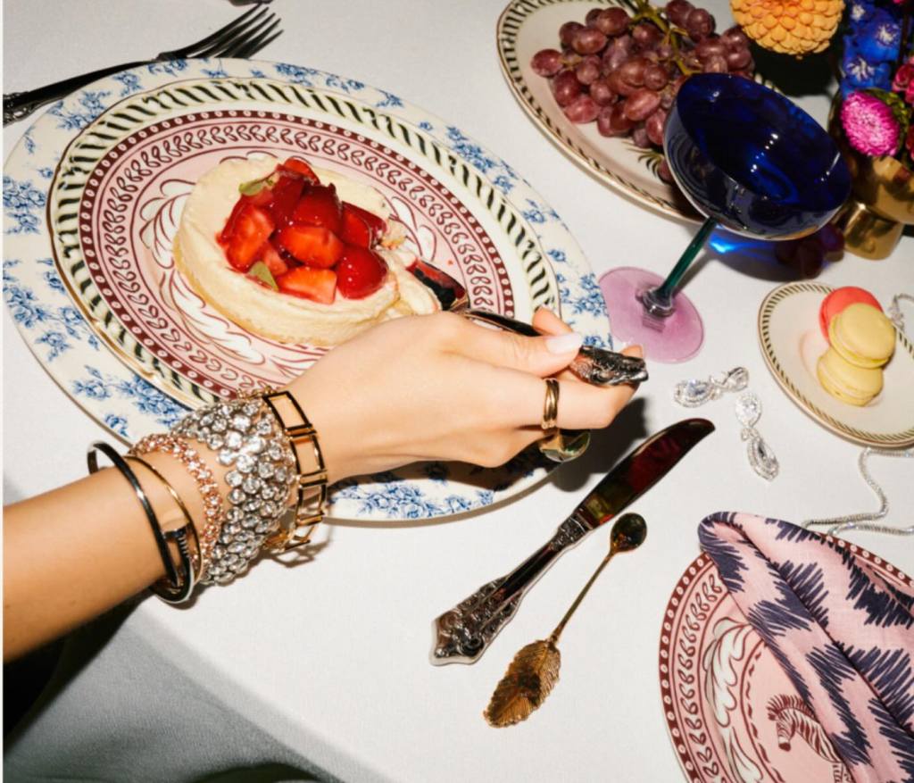 a hand draped in gold jewelry and diamonds at a dining table with fantastical culinary creations