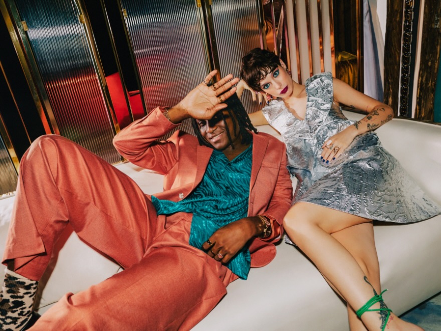 a woman in a bright silver dress sitting next to a man in an orange suit on a white couch