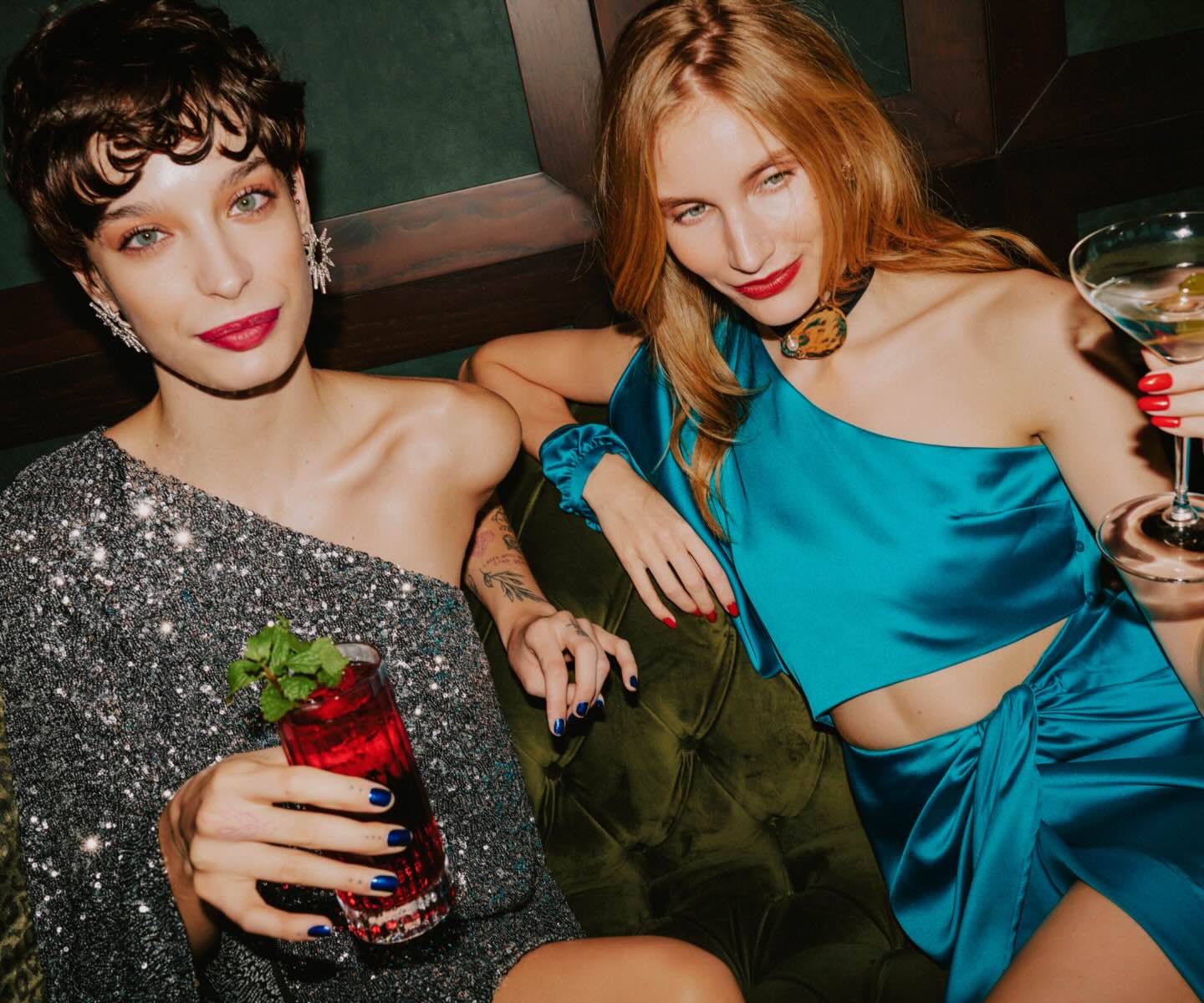 two women elegantly dressed sipping on cocktails while sitting on a velvet green couch