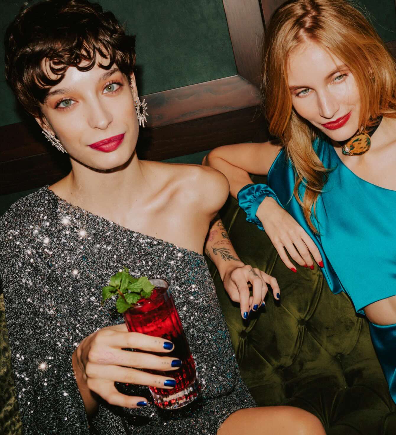 two women elegantly dressed sipping on cocktails while sitting on a velvet green couch