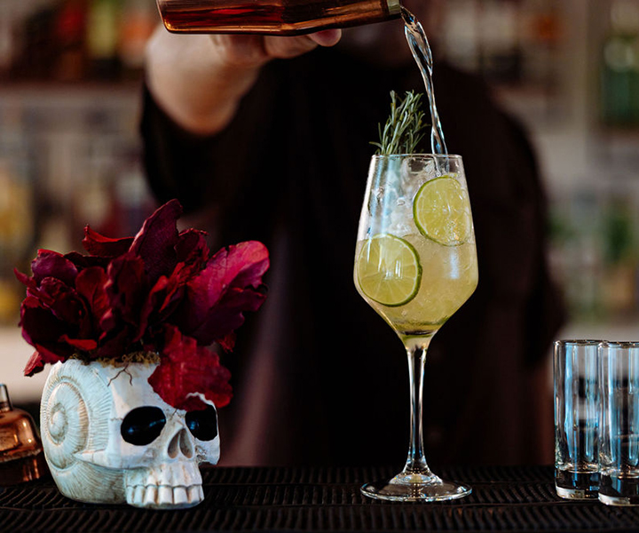 a margarita being poured into a glass by a bartender, with a skull planter holding red flowers next to it
