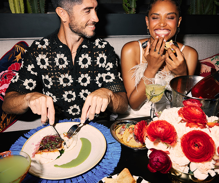 A couple laughing over a colorful table filled with flowers, Mexican food, cocktails and a bottle of pink champagne